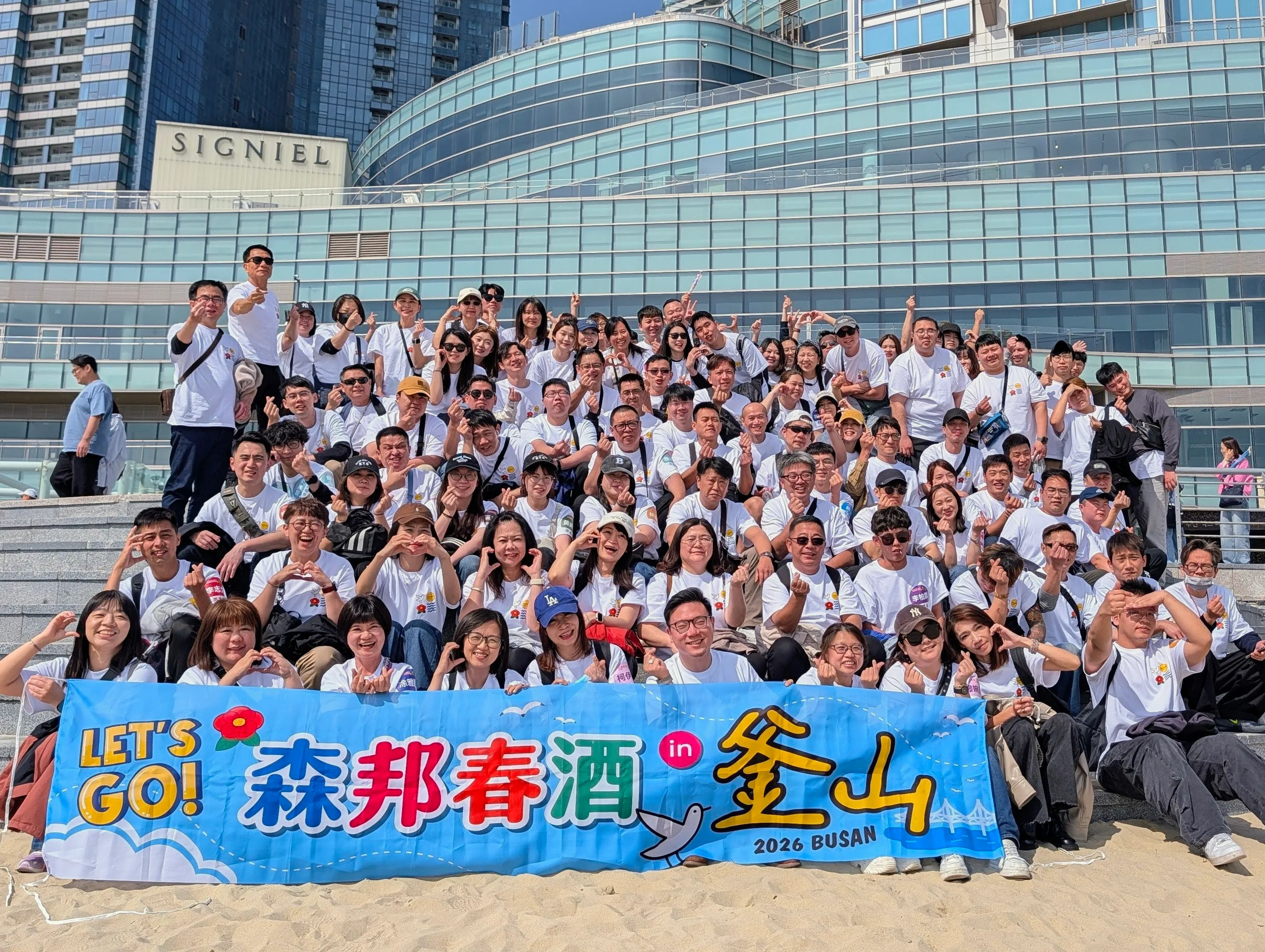 森邦集團 2026 春酒釜山合照：約一百餘名員工在海雲台沙灘前合照，全員穿白色 T-shirt